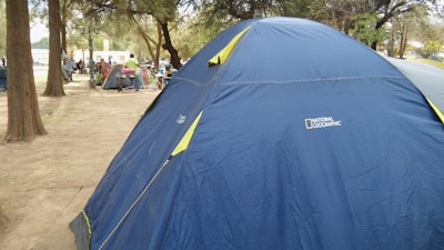 A large blue tent with yellow trim is set up in a wooded area. It is branded with the National Geographic logo. Nearby, there are other smaller tents and some people engaged in activities. The ground is covered with light brown dirt, and tall trees provide shade.