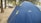 A large blue tent with yellow trim is set up in a wooded area. It is branded with the National Geographic logo. Nearby, there are other smaller tents and some people engaged in activities. The ground is covered with light brown dirt, and tall trees provide shade.