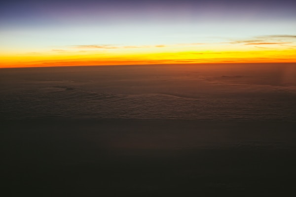 A stunning horizon at sunset with vibrant orange and yellow hues blending into the darkening sky above. The lower part of the image shows a sea of clouds, lending a serene and peaceful atmosphere.