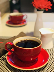 A warm cup of coffee steaming on a red and white checkered tablecloth with a baseball cap in the background.