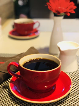 A warm cup of coffee steaming on a red and white checkered tablecloth with a baseball cap in the background.