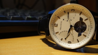 A wristwatch with a beige dial and black leather strap, featuring a seagull design and multiple subdials. The timepiece is placed on a wooden surface with an out-of-focus keyboard visible in the background.