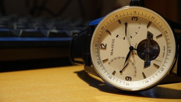 A wristwatch with a beige dial and black leather strap, featuring a seagull design and multiple subdials. The timepiece is placed on a wooden surface with an out-of-focus keyboard visible in the background.