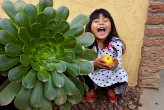 A young girl with long dark hair is smiling brightly while holding an orange. She is wearing a white dress with black polka dots and red shoes. Beside her is a large green succulent plant with thick, fleshy leaves. The background includes a yellow stucco wall and some brickwork.