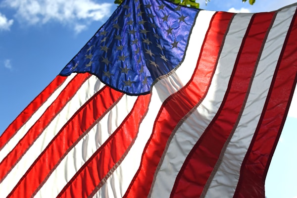 The image displays the American flag waving with the blue sky and white clouds in the background. The flag features red and white stripes alongside a blue field with white stars.