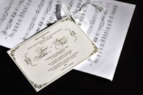 The image depicts a decorative wedding invitation card placed over sheets of music notation. The invitation is beautifully designed with elegant script and ornate borders. Attached to the invitation is a delicate white ribbon.