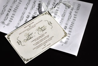 The image depicts a decorative wedding invitation card placed over sheets of music notation. The invitation is beautifully designed with elegant script and ornate borders. Attached to the invitation is a delicate white ribbon.