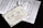 The image depicts a decorative wedding invitation card placed over sheets of music notation. The invitation is beautifully designed with elegant script and ornate borders. Attached to the invitation is a delicate white ribbon.