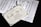 The image depicts a decorative wedding invitation card placed over sheets of music notation. The invitation is beautifully designed with elegant script and ornate borders. Attached to the invitation is a delicate white ribbon.