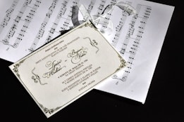 The image depicts a decorative wedding invitation card placed over sheets of music notation. The invitation is beautifully designed with elegant script and ornate borders. Attached to the invitation is a delicate white ribbon.
