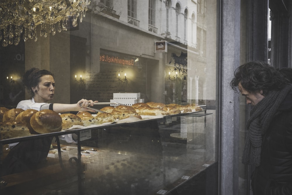 man facing storefront in front of woman holding thong picking bread