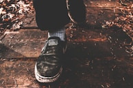 A close-up of a person's foot wearing a black shoe stepping on a wooden surface. The shoe has visible wear and features a label with text. The ground is covered with dry leaves and small twigs, giving a rustic appearance.