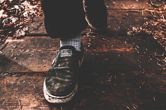 A close-up of a person's foot wearing a black shoe stepping on a wooden surface. The shoe has visible wear and features a label with text. The ground is covered with dry leaves and small twigs, giving a rustic appearance.