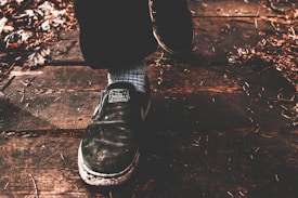 A close-up of a person's foot wearing a black shoe stepping on a wooden surface. The shoe has visible wear and features a label with text. The ground is covered with dry leaves and small twigs, giving a rustic appearance.