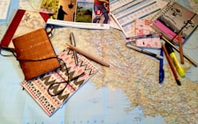 A large map is spread out on a table, surrounded by various stationery items including notebooks, pens, pencils, a highlighter, and brochures or travel-related pamphlets. The scene suggests planning, mapping out a journey, or organizing travel details.