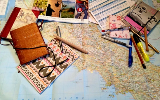 A large map is spread out on a table, surrounded by various stationery items including notebooks, pens, pencils, a highlighter, and brochures or travel-related pamphlets. The scene suggests planning, mapping out a journey, or organizing travel details.