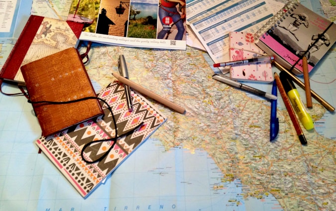 A large map is spread out on a table, surrounded by various stationery items including notebooks, pens, pencils, a highlighter, and brochures or travel-related pamphlets. The scene suggests planning, mapping out a journey, or organizing travel details.
