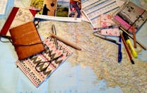 A large map is spread out on a table, surrounded by various stationery items including notebooks, pens, pencils, a highlighter, and brochures or travel-related pamphlets. The scene suggests planning, mapping out a journey, or organizing travel details.