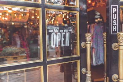 A storefront door with a glass window that has an 'OPEN' sign hanging on it. The door features gold-trimmed frames and ornate handles. Through the glass, a warmly lit interior is visible with hanging clothes and decorative lights.