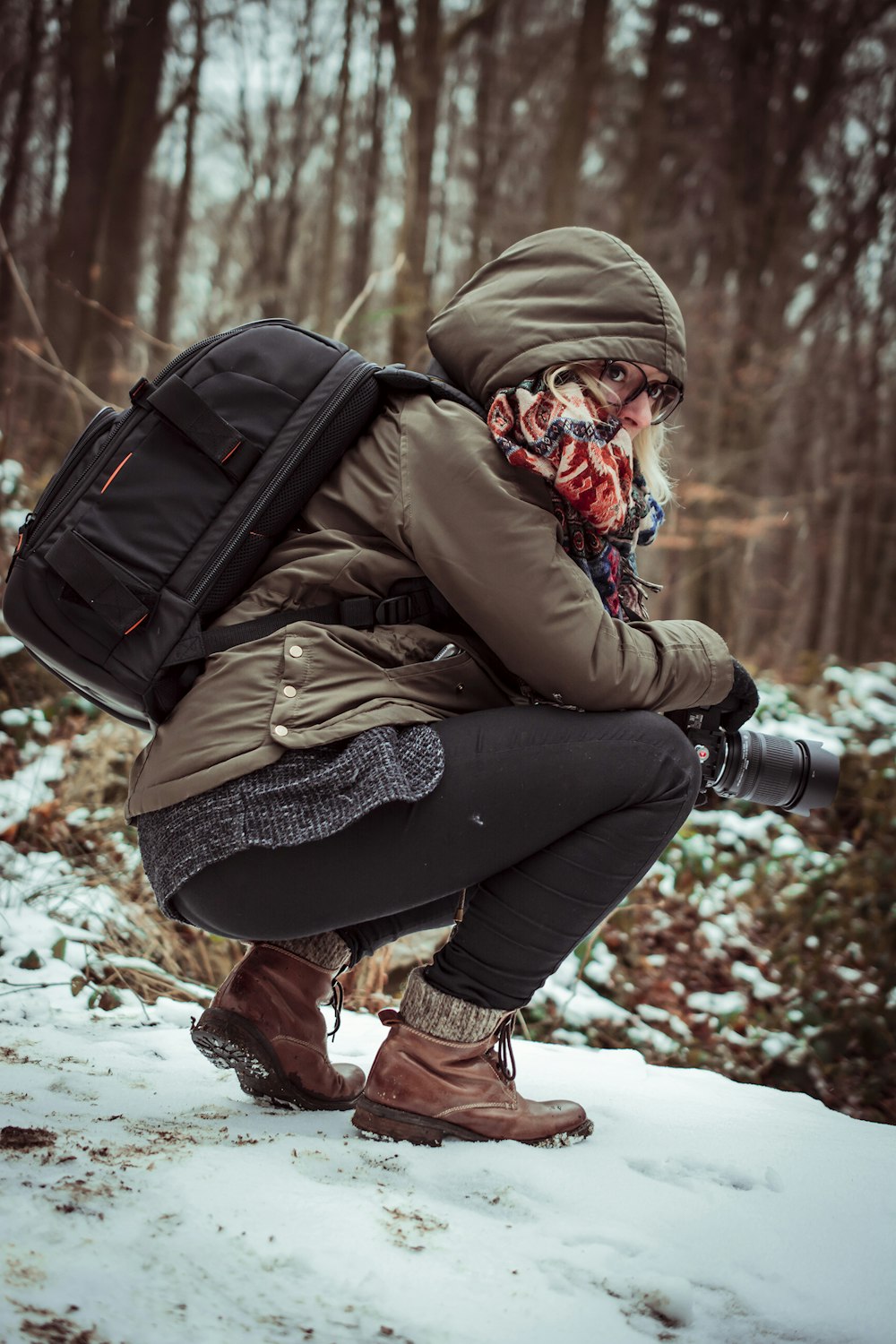 日中、雪に覆われた地面にしゃがみながらカメラを構える茶色のフード付きジャケットを着た女性