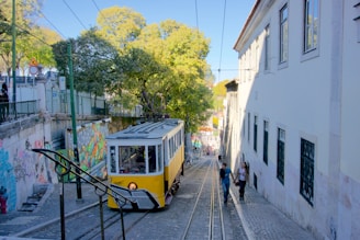 A yellow tram is ascending a cobblestone street lined with buildings and graffiti-covered walls. There are two people walking alongside the tram tracks, and trees with green foliage are visible in the background. The scene is bathed in daylight, creating a bright and lively atmosphere.