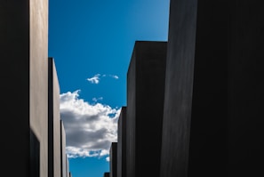 Tall, monochromatic concrete blocks create a geometric pattern against a clear blue sky with a few scattered clouds.