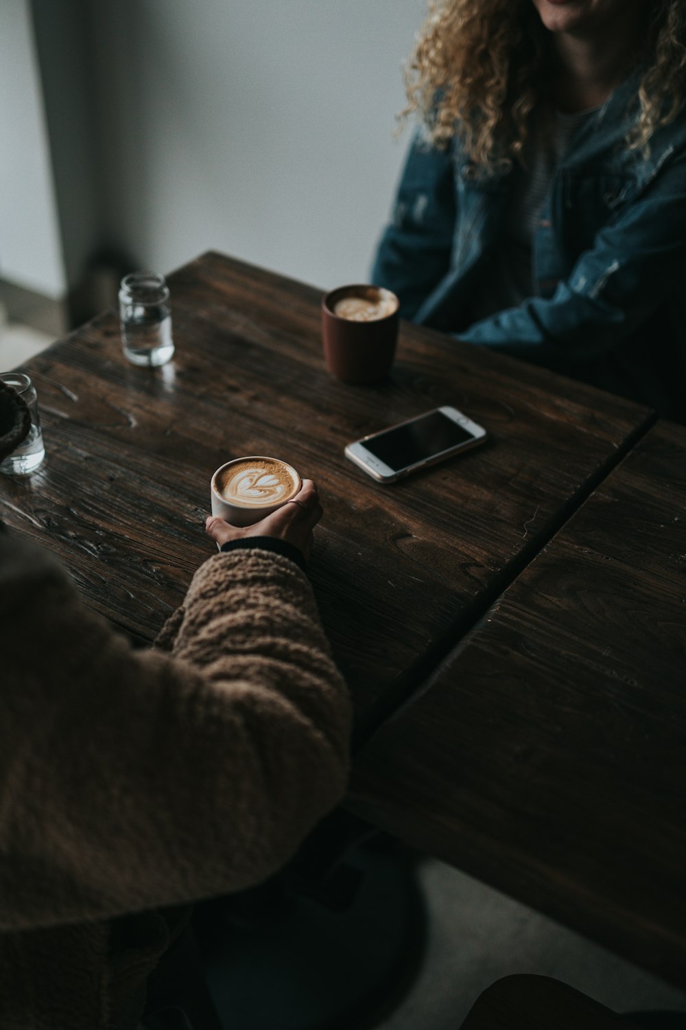 갈색 스웨터를 입은 사람이 방 안의 테이블 앞에 앉아 컵을 들고 있습니다.