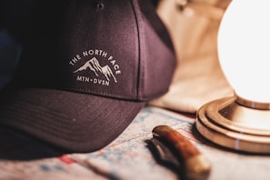 A dark-colored cap with a mountain emblem and text placed on a table. Beside it is a lit lamp with a metallic base, illuminating the surroundings with a warm glow. A map is spread under the objects, partially covered by a folded knife with a wooden handle.