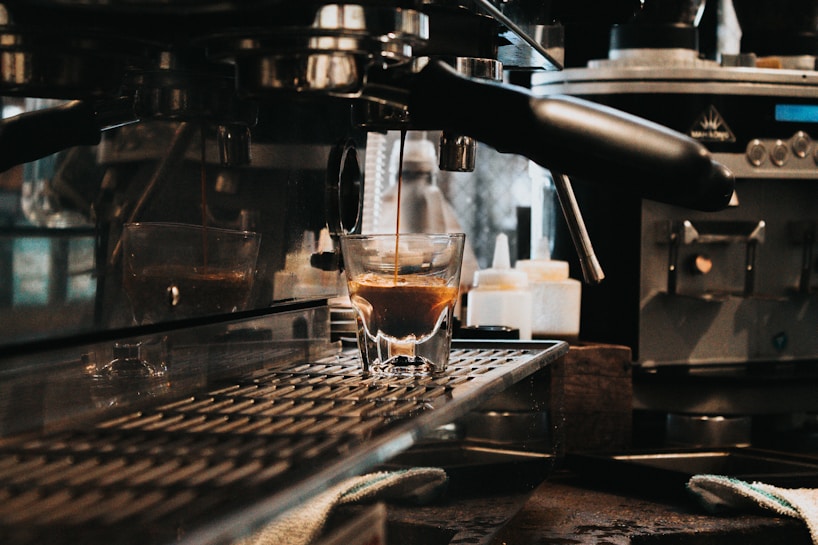A close-up view of an espresso machine in operation, with a single shot of espresso being poured into a small glass. The surrounding area includes various coffee machine components and accessories, indicating a café or kitchen setting.