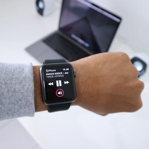A person wearing a smartwatch displaying a music player app. The screen shows a song titled 'KNOCK KNOCK' by TWICE. A laptop and a small speaker are visible in the background on a white desk.