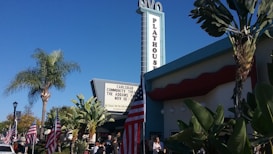 A theater building with a large vertical 'Playhouse' sign and a marquee advertising a performance. American flags are displayed prominently, and the scene is framed by palm trees under a clear blue sky. People are gathered around the entrance.
