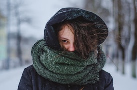 A person is bundled up in winter clothing, with a large dark green scarf wrapped around their neck and a hood pulled up over their head. Their hair appears windswept, and snowflakes are visible on the attire. The background is a snowy landscape with blurry trees.