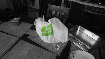 A dining table is set with several items, including a white shopping bag with green writing, a glass tumbler, a metal roasting pan, and a plate with a leaf design. Several placemats are laid out on the table, and a pack of playing cards can be seen on the side.