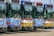 Four city buses are parked side by side in a row, each displaying different colors such as blue, red, and yellow. They are equipped with bike racks on their fronts and show route numbers above the windshield.