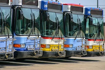 Four city buses are parked side by side in a row, each displaying different colors such as blue, red, and yellow. They are equipped with bike racks on their fronts and show route numbers above the windshield.