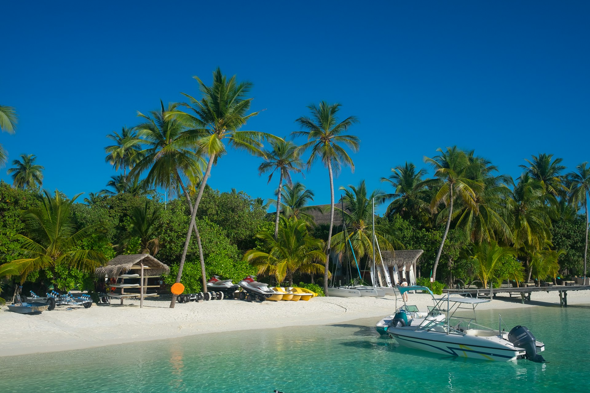 A tropical beach scene featuring clear aqua water, white sandy shore, and lush green palm trees. Jet skis are lined up near a small wooden hut. A motorboat is anchored by the shore, with bright blue skies overhead.