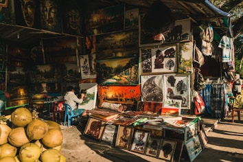 A street market stall displays a variety of vibrant paintings and artworks. The artwork includes landscape paintings, portraits, and intricate cut-out designs. A person is seated and actively painting on a canvas. There are stacks of coconuts in the foreground, adding a tropical element to the scene. The overall ambiance is colorful and bustling, with other stalls and items such as fabrics and bags surrounding the main focus.