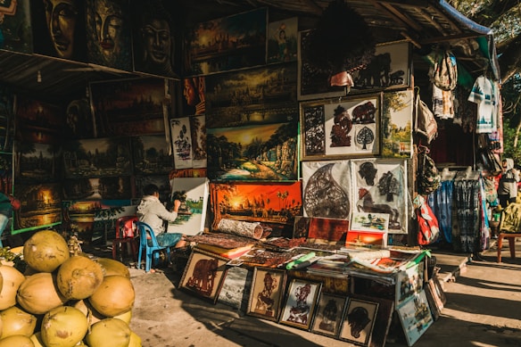 A street market stall displays a variety of vibrant paintings and artworks. The artwork includes landscape paintings, portraits, and intricate cut-out designs. A person is seated and actively painting on a canvas. There are stacks of coconuts in the foreground, adding a tropical element to the scene. The overall ambiance is colorful and bustling, with other stalls and items such as fabrics and bags surrounding the main focus.