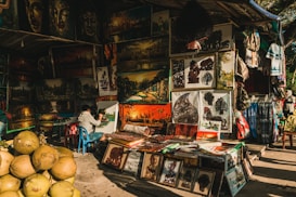 A street market stall displays a variety of vibrant paintings and artworks. The artwork includes landscape paintings, portraits, and intricate cut-out designs. A person is seated and actively painting on a canvas. There are stacks of coconuts in the foreground, adding a tropical element to the scene. The overall ambiance is colorful and bustling, with other stalls and items such as fabrics and bags surrounding the main focus.