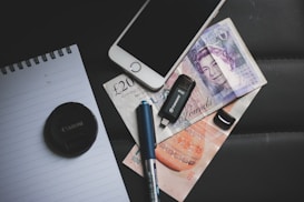 A collection of items is arranged on a dark surface, including a mobile phone, British currency notes, a USB flash drive, a black camera lens cap, a pen, and a lined notebook.