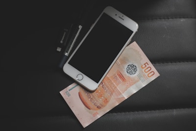 A smartphone with a blank screen is placed on top of a 500 currency note, accompanied by a USB flash drive. These items lie on a dark, textured surface.