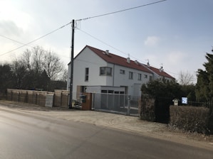 A residential building with a sloping red roof and multiple chimneys is located at the corner of a street. The building has white walls, several windows, and a gated entrance. In the foreground, a paved road runs parallel to the building. On the left side, there is a tall utility pole with hanging wires. To the side of the building, a wooden fence and some trees are visible, suggesting a suburban or semi-rural setting.