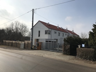 A residential building with a sloping red roof and multiple chimneys is located at the corner of a street. The building has white walls, several windows, and a gated entrance. In the foreground, a paved road runs parallel to the building. On the left side, there is a tall utility pole with hanging wires. To the side of the building, a wooden fence and some trees are visible, suggesting a suburban or semi-rural setting.