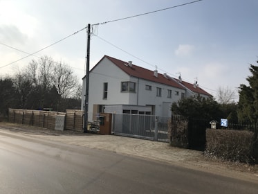 A residential building with a sloping red roof and multiple chimneys is located at the corner of a street. The building has white walls, several windows, and a gated entrance. In the foreground, a paved road runs parallel to the building. On the left side, there is a tall utility pole with hanging wires. To the side of the building, a wooden fence and some trees are visible, suggesting a suburban or semi-rural setting.