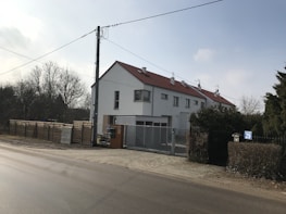 A residential building with a sloping red roof and multiple chimneys is located at the corner of a street. The building has white walls, several windows, and a gated entrance. In the foreground, a paved road runs parallel to the building. On the left side, there is a tall utility pole with hanging wires. To the side of the building, a wooden fence and some trees are visible, suggesting a suburban or semi-rural setting.