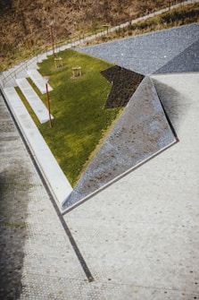 A geometric landscape design featuring a mix of materials, including a triangular patch of green grass, gravel, and white pavement. The area is bordered by a low fence, and several small trees are planted in the grass. Red lamp posts provide a pop of color against the neutral tones.