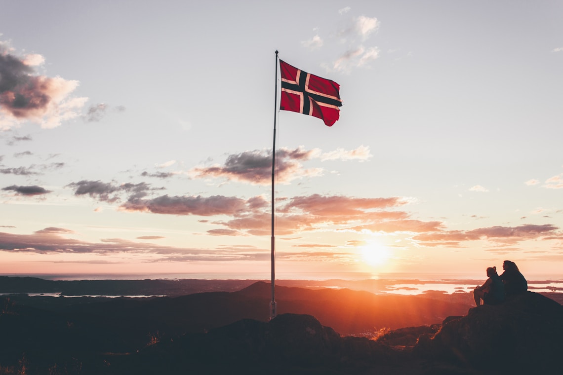 norway flag at full mast on cliff with two people sat beside it at sunset
