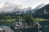 A serene mountain landscape with a clear lake reflecting the surrounding greenery and tall rocky peaks. A person is standing on a rock in the middle of the lake, practicing yoga amidst lush vegetation.