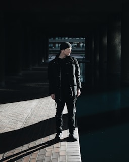 A person wearing dark clothing stands on a paved surface, partially illuminated by a strong light source creating distinct shadows. The background consists of a dark, architectural space with visible pillars and a body of water nearby. The person is looking towards the right, adding a contemplative element to the scene.
