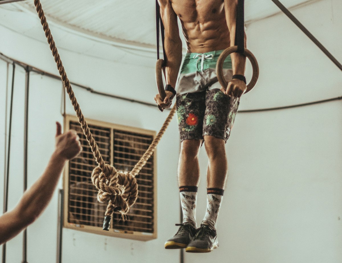 Athlète suspendu aux anneaux de gymnastique à côté d'une corde à grimper, un coach l'encourage pouce levé